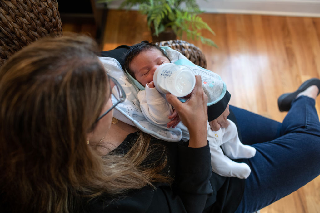 Woman with brown hair feeds baby with a bottle of milk. The baby is infant sized and wearing a large blue bib.